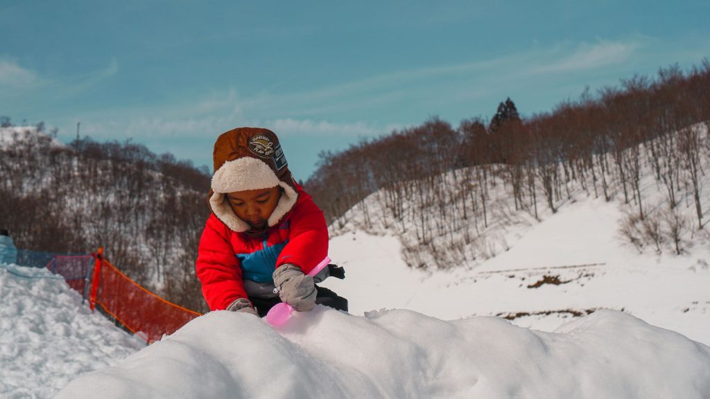 A child in a red jacket plays in the snow amidst a scenic winter mountain landscape.