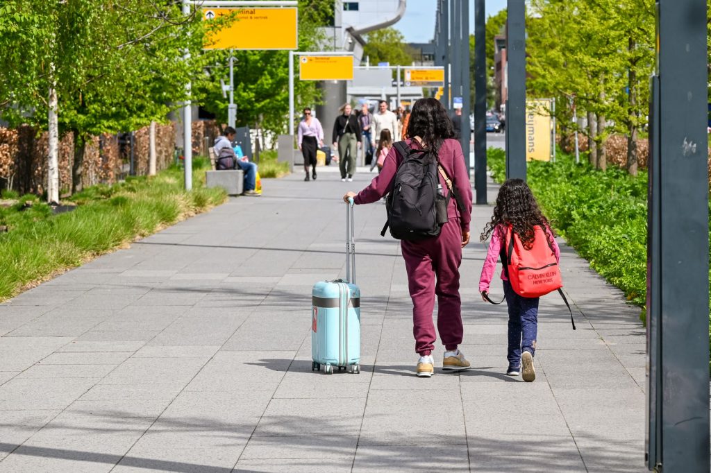 A mother and daughter walking with luggage outside Eindhoven Airport on a sunny day.