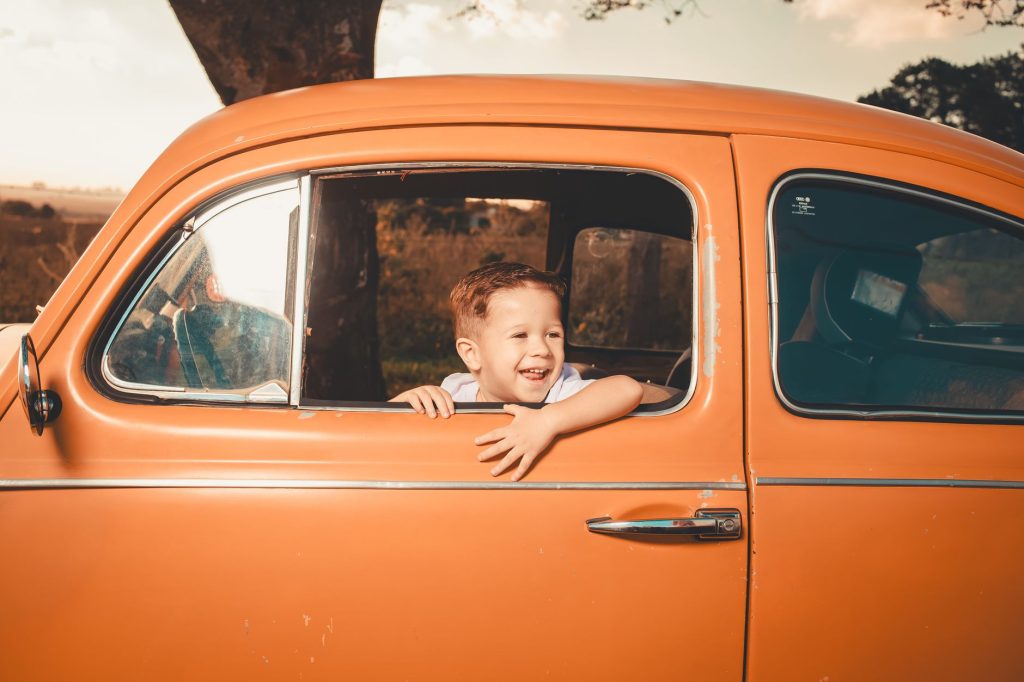 A joyful child peeks out of a window of a vintage orange car, enjoying a sunny day outdoors.