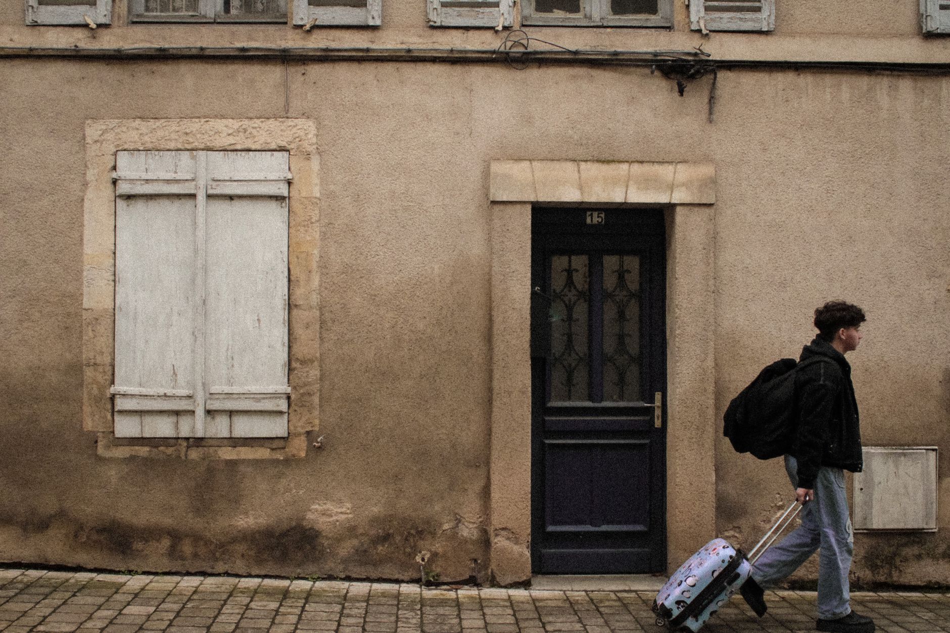 Man walking with luggage past a vintage building exterior with wooden shutters.