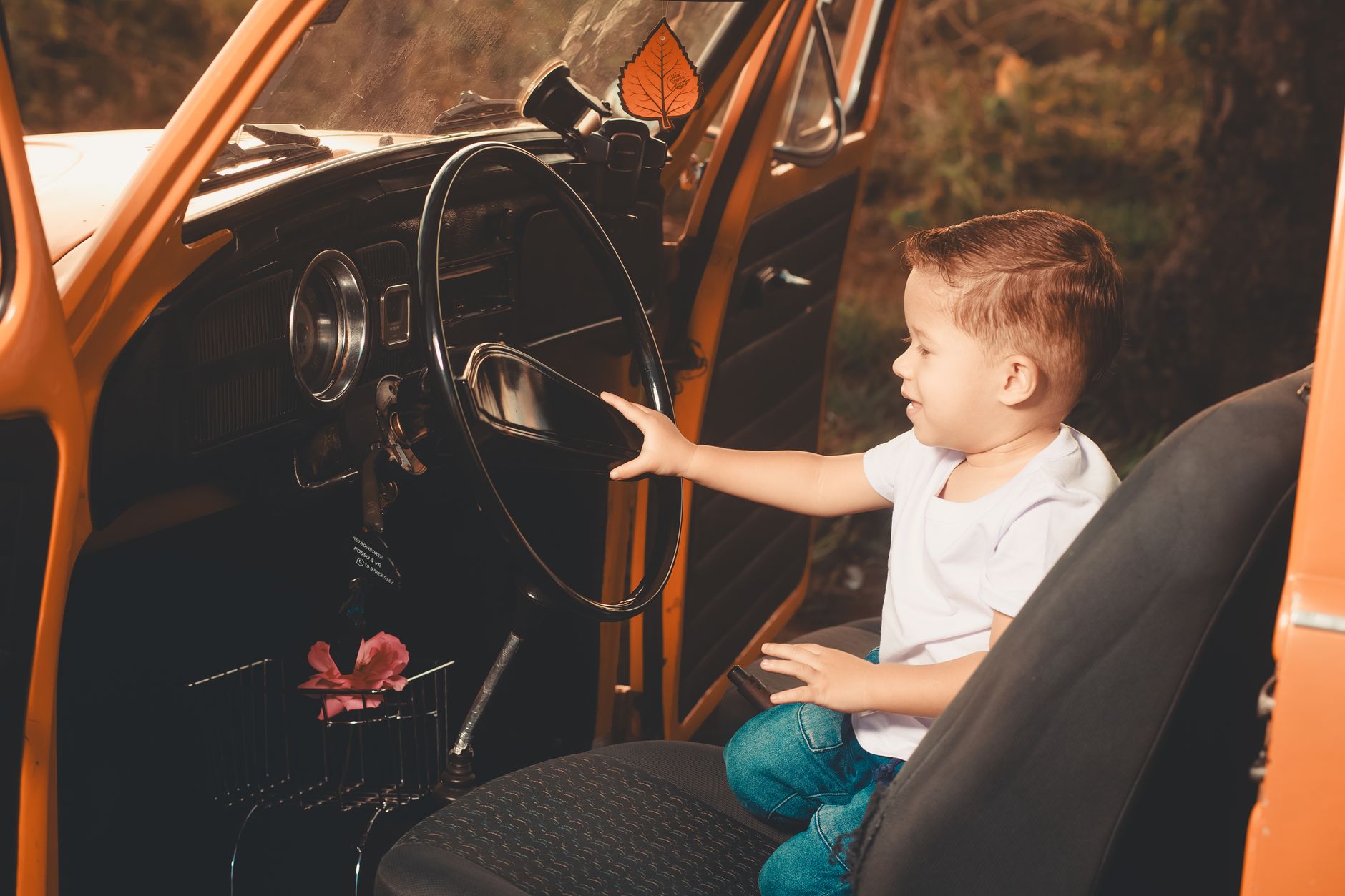 A young boy sits in a classic car, playfully holding the steering wheel while smiling.