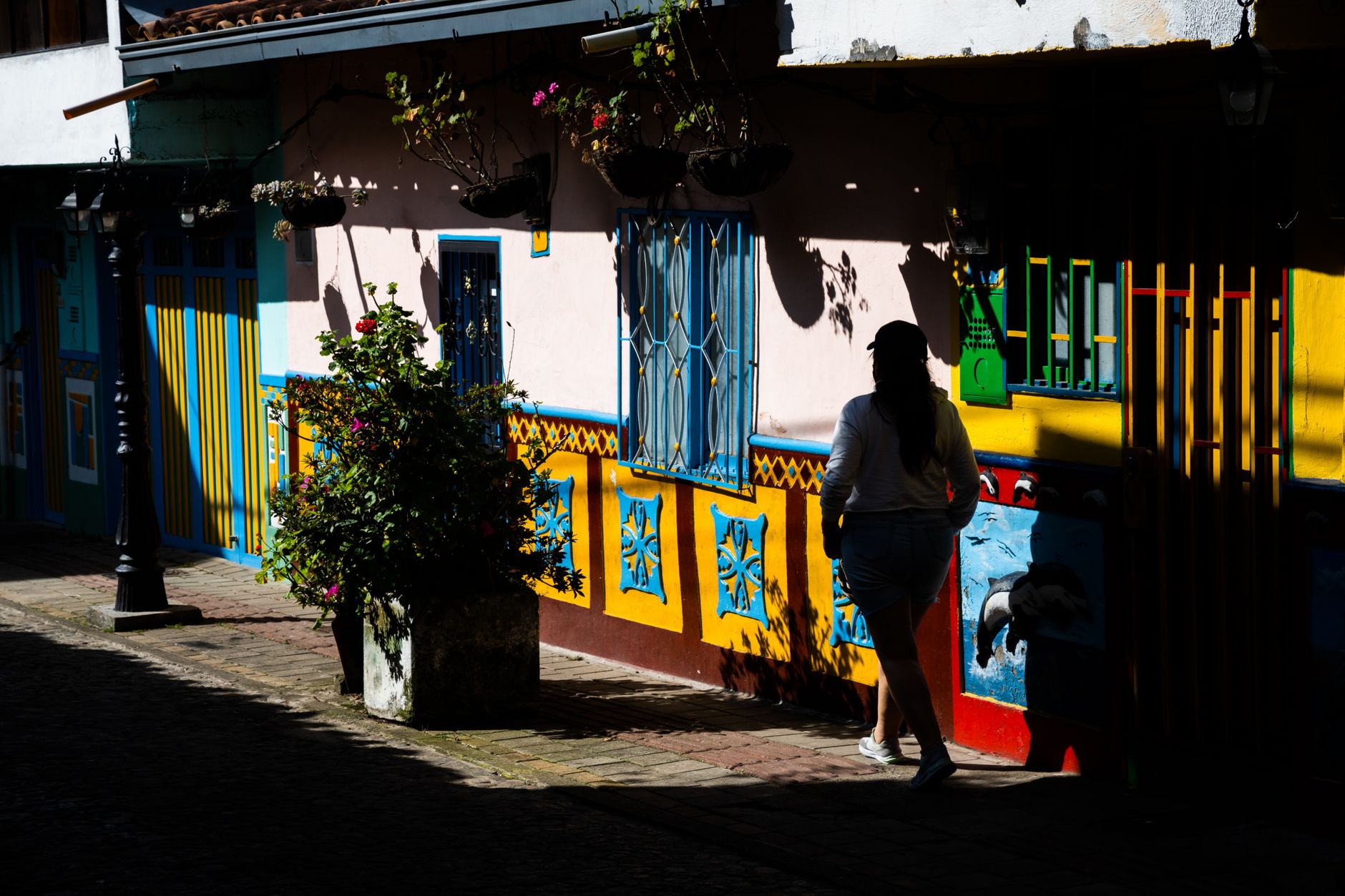 A woman walks along a vibrant street in Guatapé, Colombia, featuring colorful buildings and hanging plants.