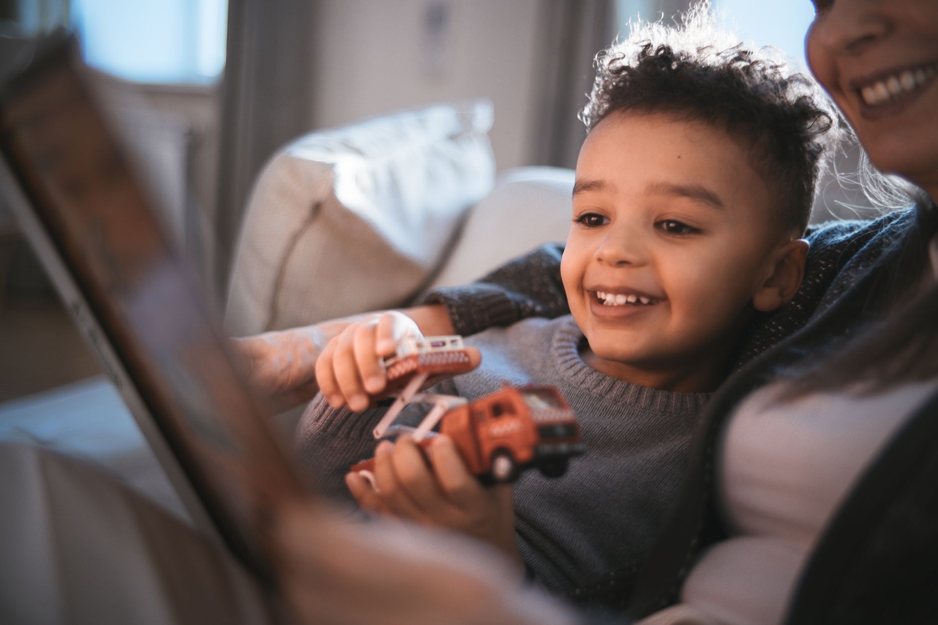 A joyful moment of a mother and son bonding over a book and toy cars indoors.