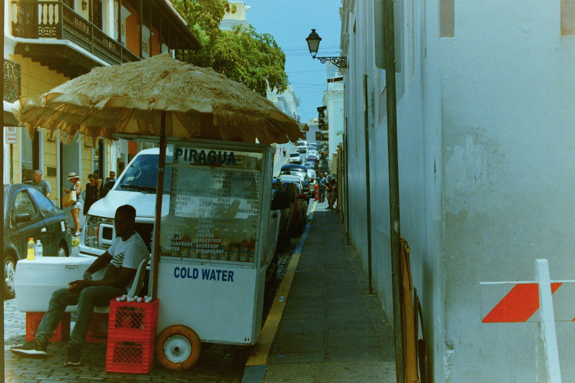 A vibrant street vendor selling piragua, under a straw umbrella in an urban environment, offering cold water.