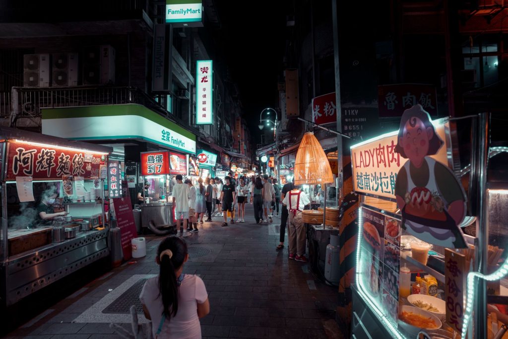 Bustling night market with diverse street food stalls and people shopping under vibrant lights.