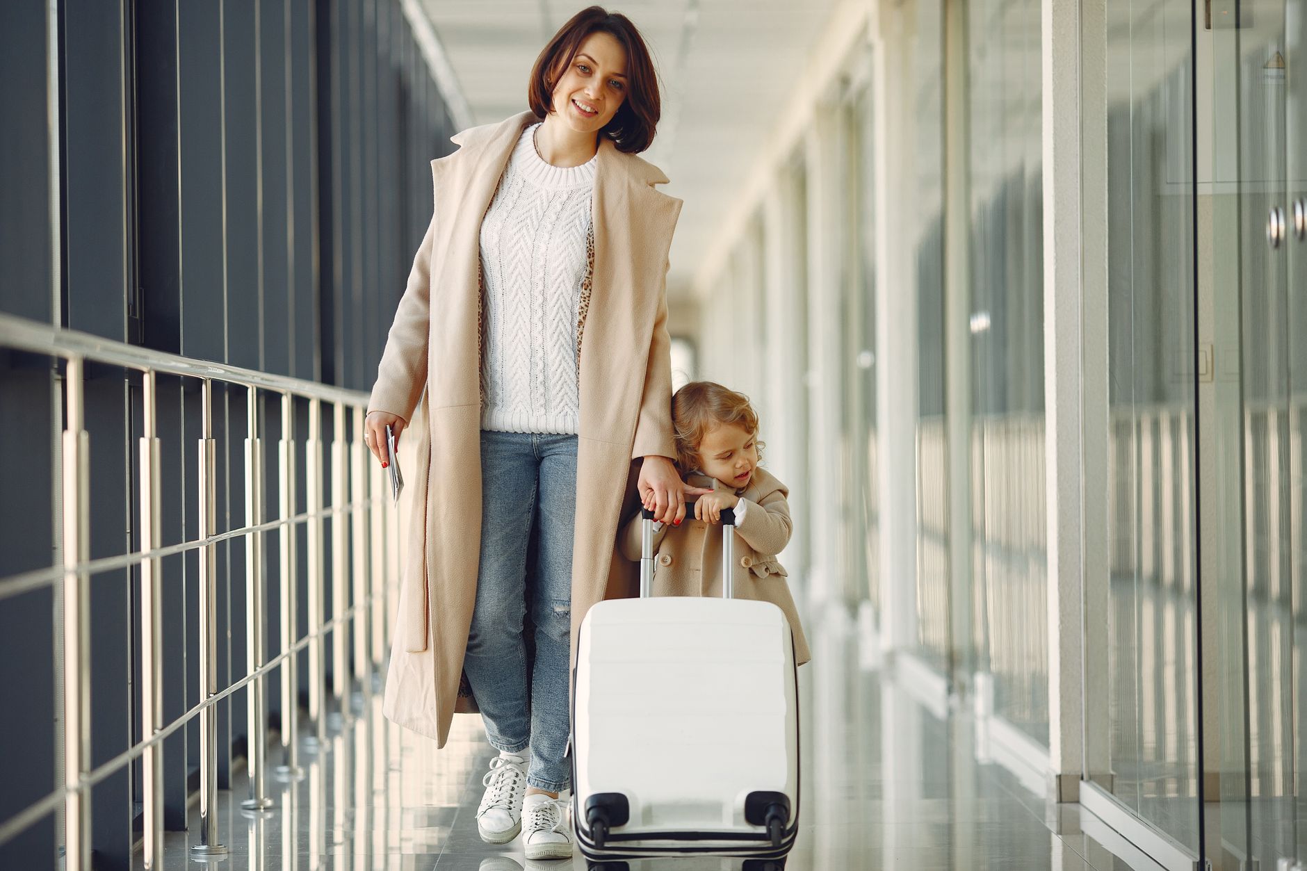 Full length of content woman with daughter in jeans and beige coats carrying white suitcase while walking along modern airport airbridge
