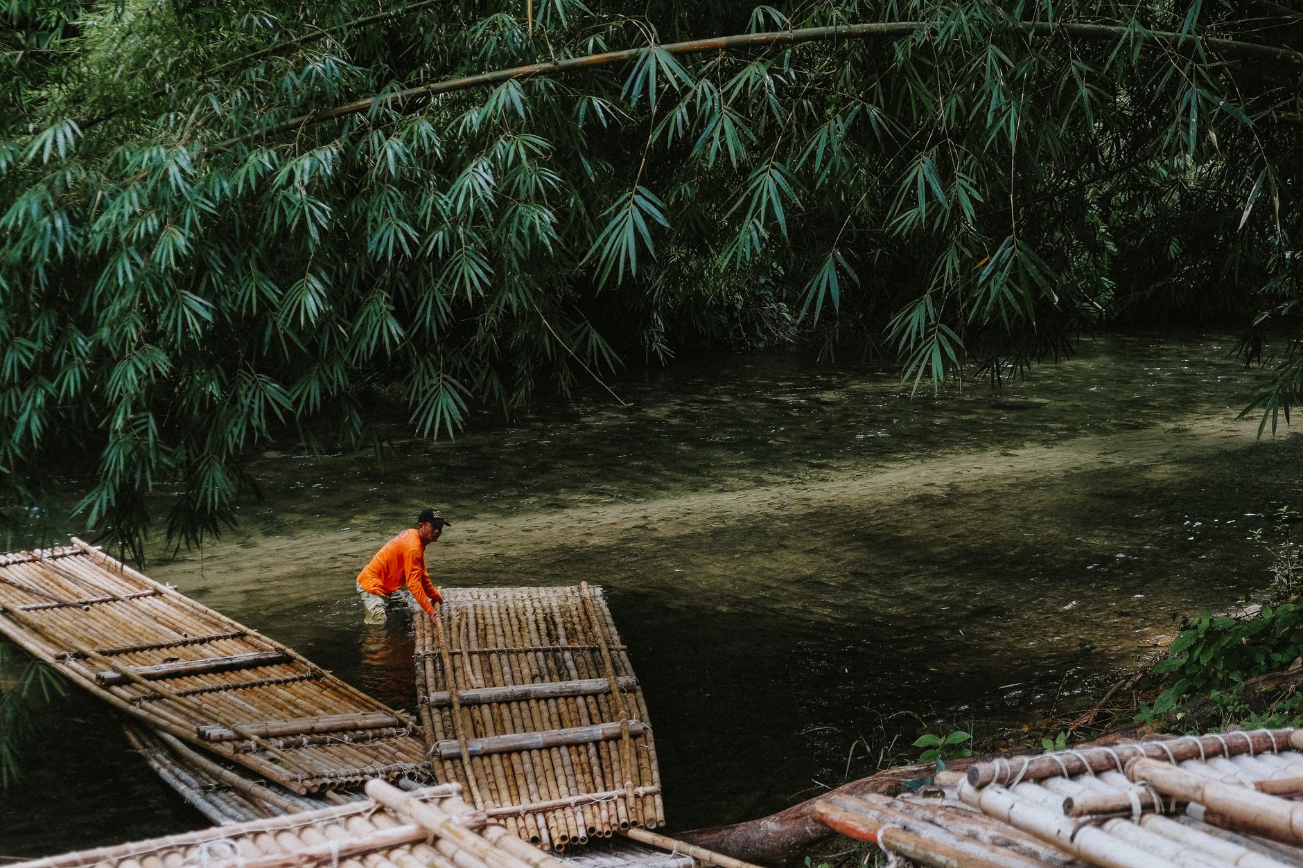 A lone man navigates a bamboo raft under lush green bamboo trees on a calm tropical river.