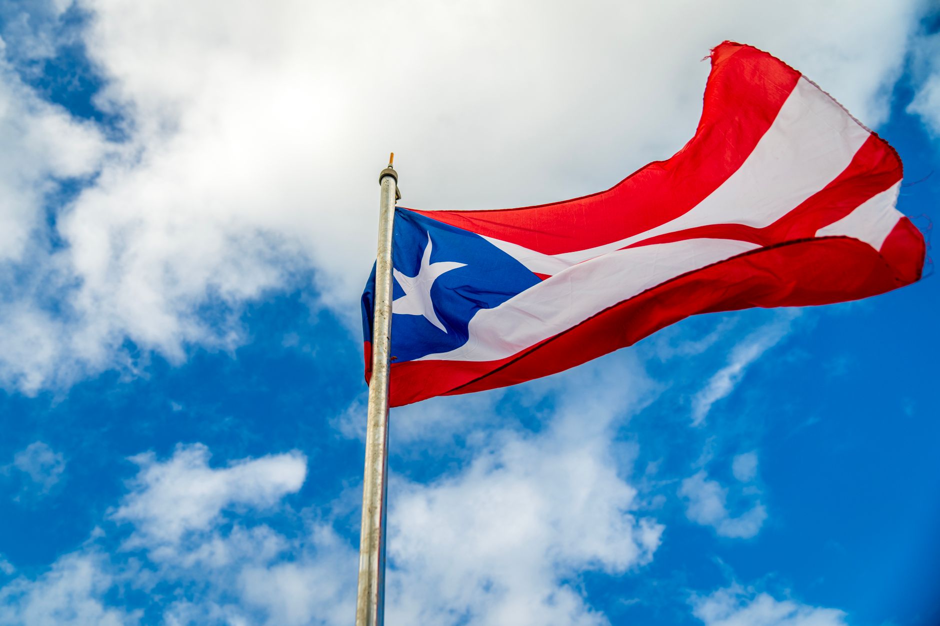 A vibrant Puerto Rican flag waving on a flagpole against a bright blue sky with clouds.