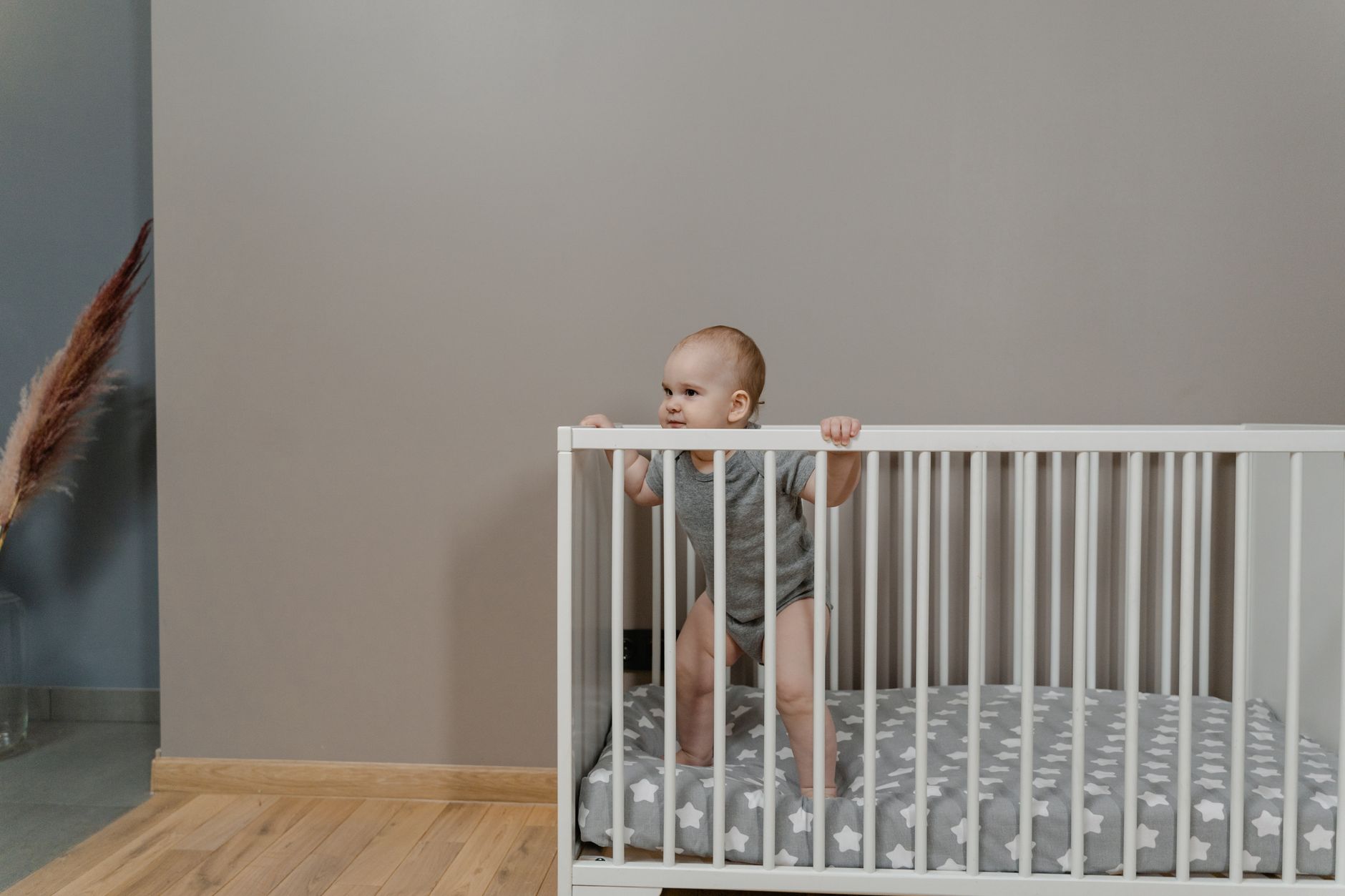 A cute baby in a grey onesie stands in a white crib indoors.