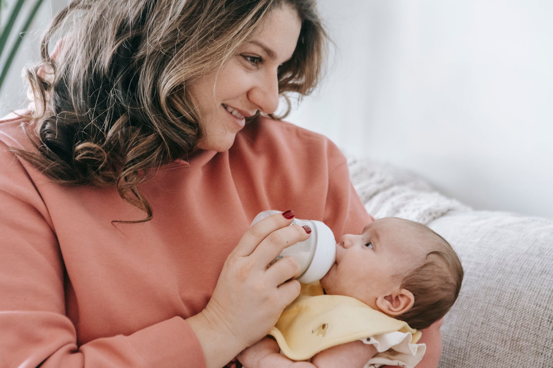 Optimistic crop mother with curly hair feeding infant baby with bottle while sitting on comfortable couch in light living room at home