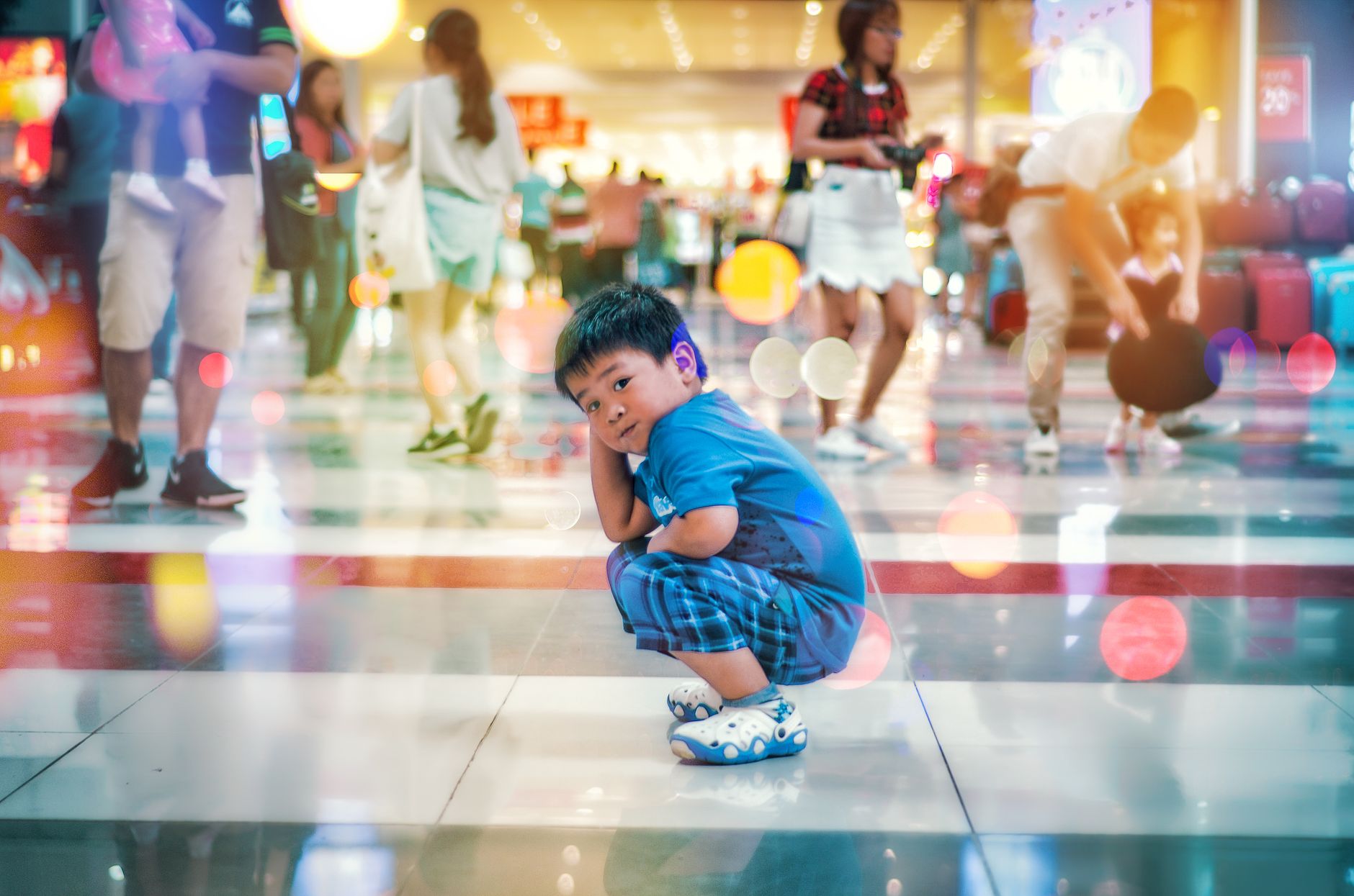 A young boy in a blue outfit sits on the floor in a colorful, bustling mall setting.