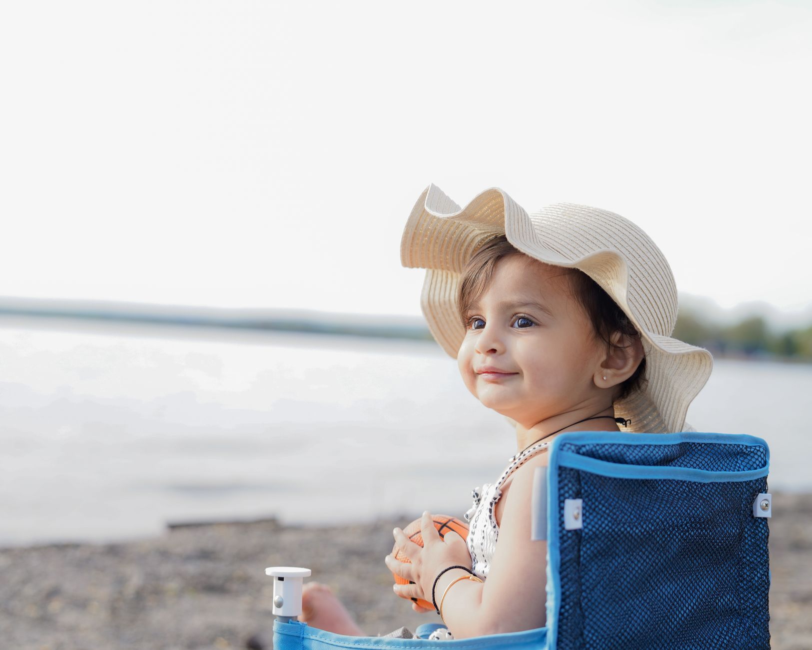 Cute baby girl with hat sitting by the beach, enjoying a serene day outdoors.