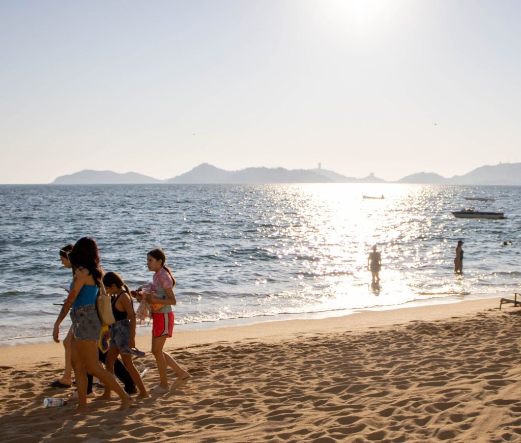 Adults and children enjoying a sunny day at the beach in Acapulco, with the sea and islands in view.