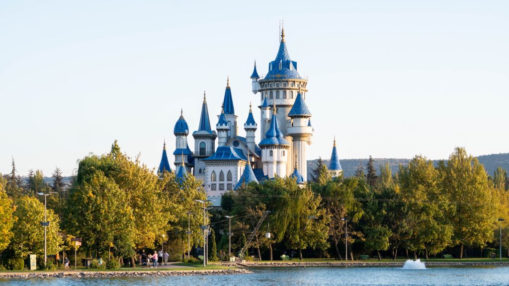 Magical fairy tale castle with blue spires surrounded by lush park and tranquil water.