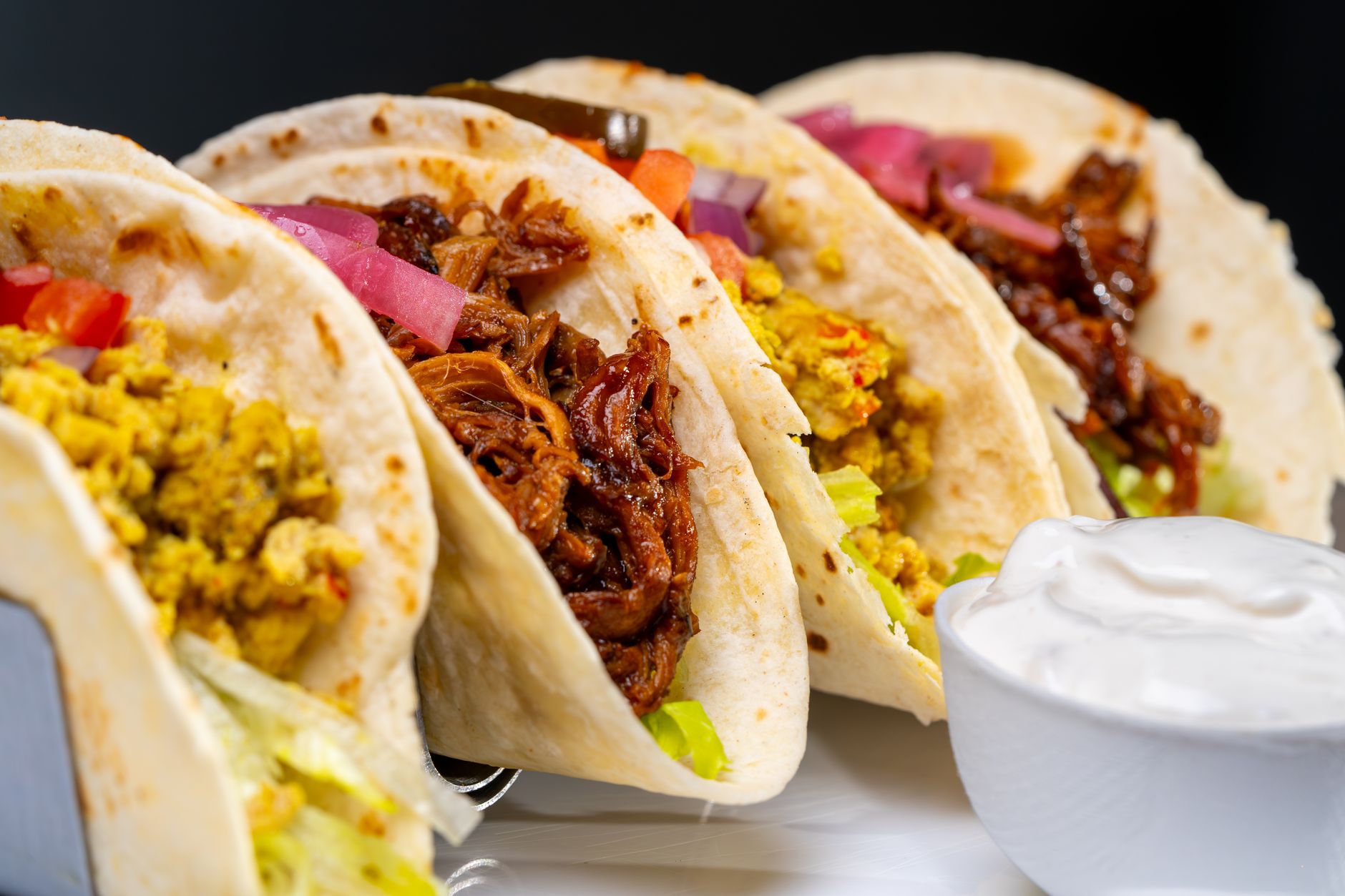 A variety of tacos with toppings and a side of creamy sauce.