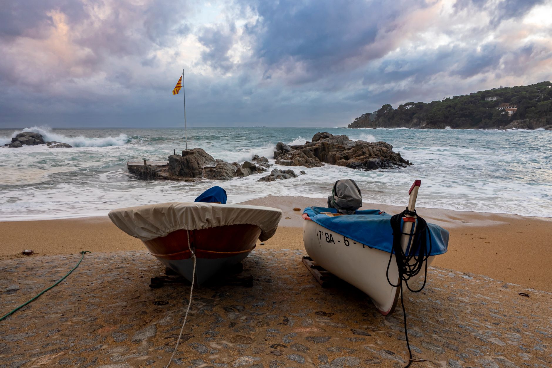 Two boats on a sandy beach in Calella de Palafrugell with waves and rocks in the background.
