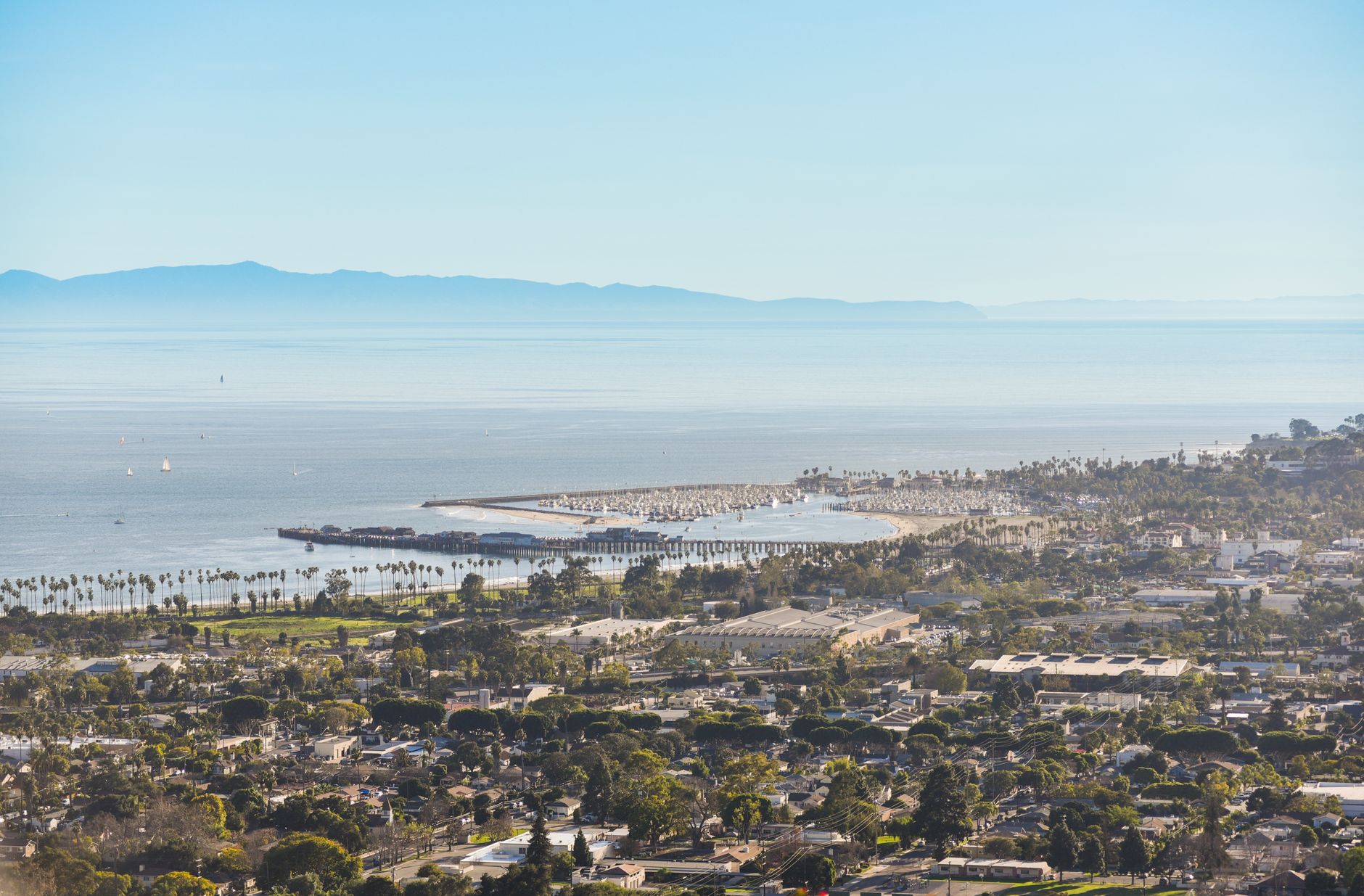 Aerial view of a scenic coastal city with ocean, beach, and mountains under a bright blue sky.