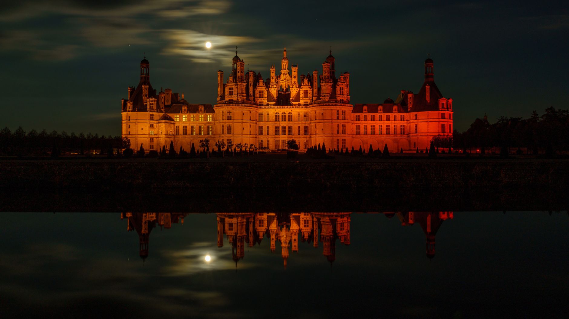 Night view of Château de Chambord reflecting in water under a full moon.