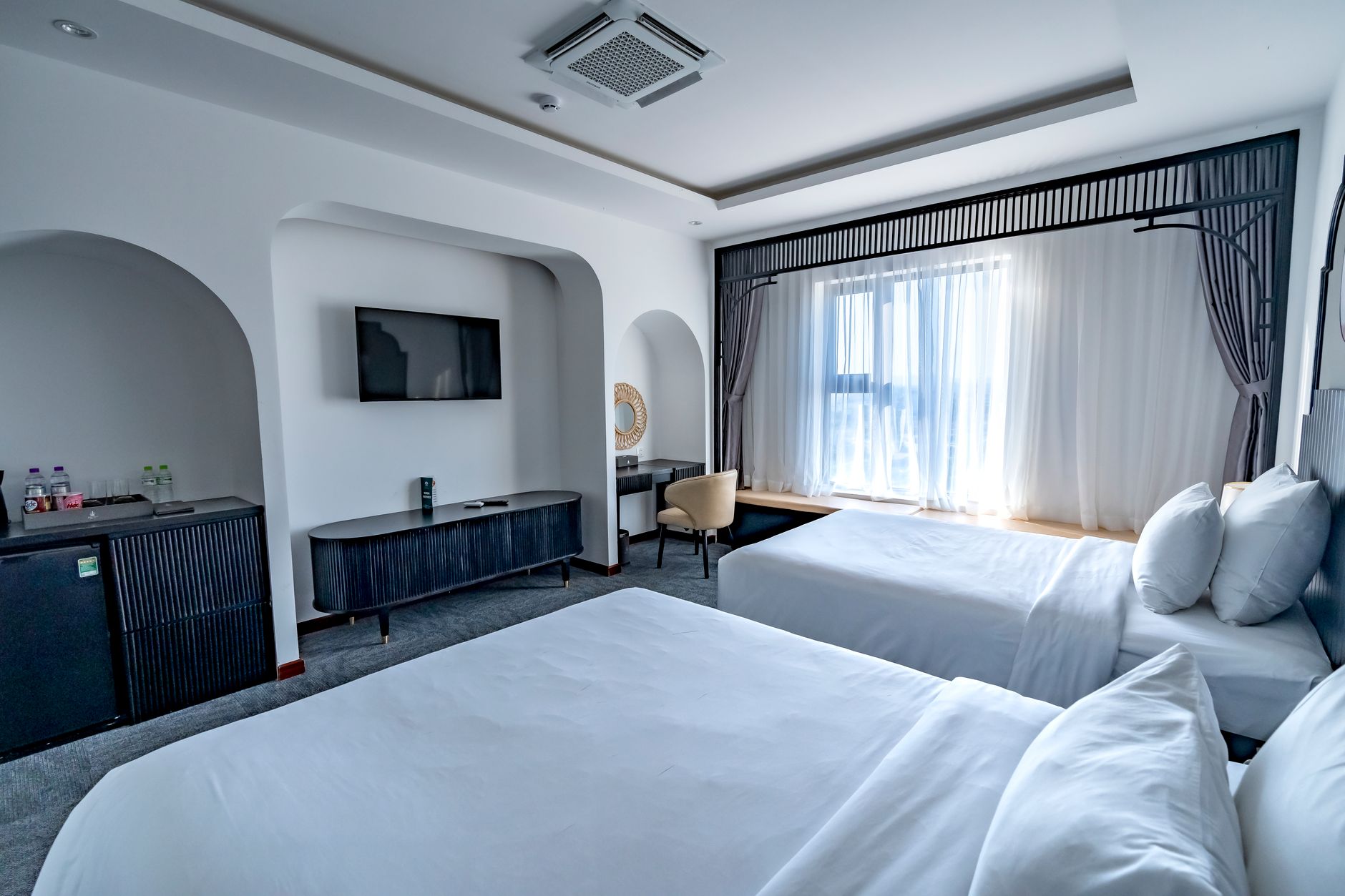 Spacious modern hotel room featuring two beds, a TV, and ample natural light through sheer curtains.