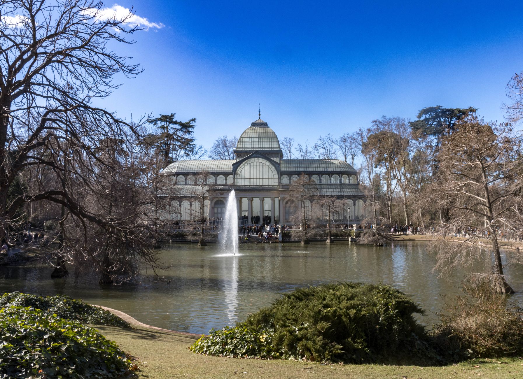 The iconic Crystal Palace in Retiro Park, Madrid, with a tranquil pond and fountain under a clear sky.