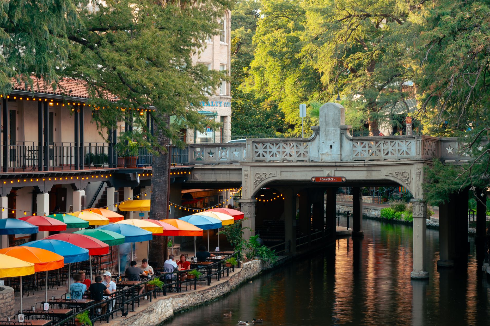 Colorful outdoor dining along the San Antonio Riverwalk with lush greenery and iconic bridge.