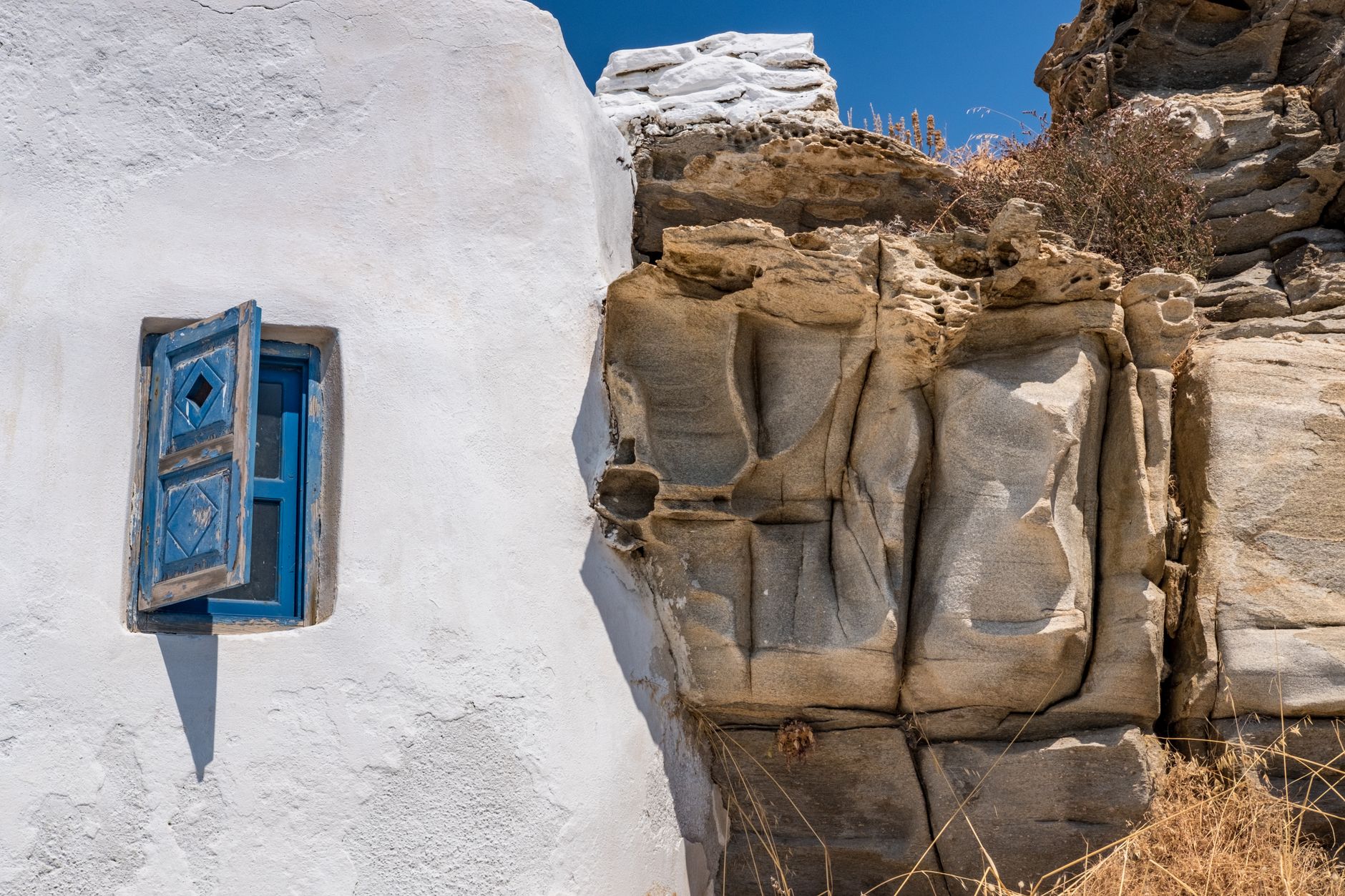 Charming rustic window in Paros, Greece, with a whitewashed wall and rock formations under a clear sky.