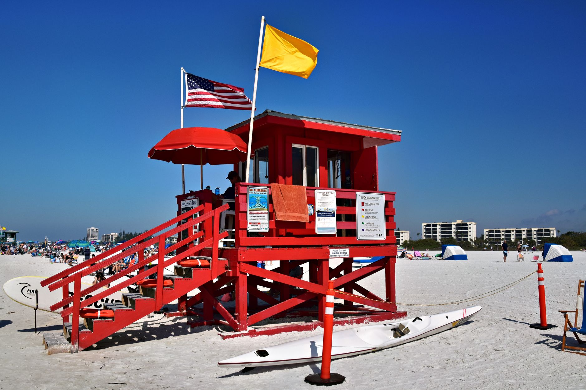 Bright red lifeguard tower with flags on a busy, sunlit beach, perfect for vacation scenery.